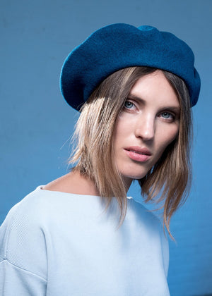 A person wearing a wool blue beret with a white sweater on a blue background 