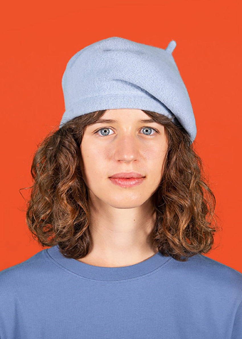A person wearing a light blue wool conical beret with a blue sweater on a orange background 