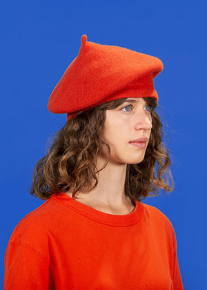 A person wearing a orange wool conical beret with matching sweater on a blue background 