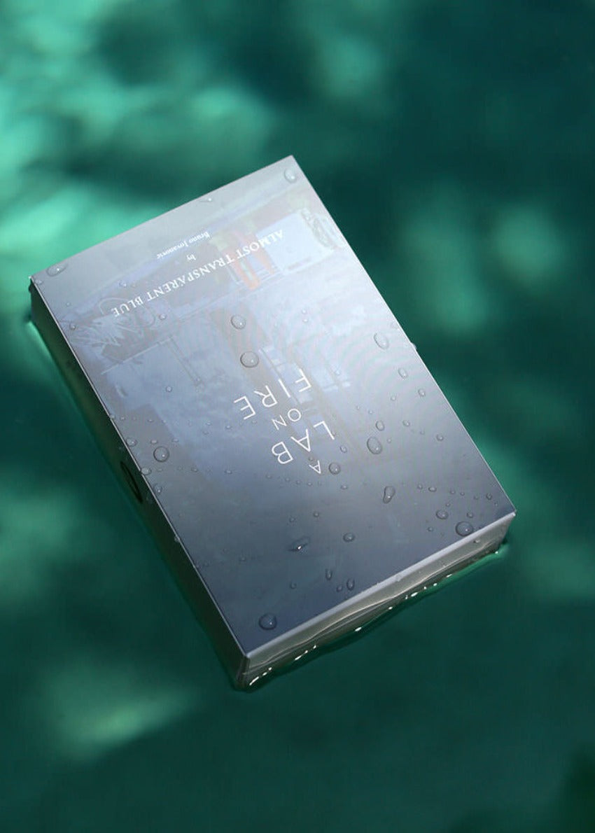 A white bottle of 'Almost Transparent Blue' fragrance with a black cap and a label that has a flame graphic and the text 'A Lab on Fire'.